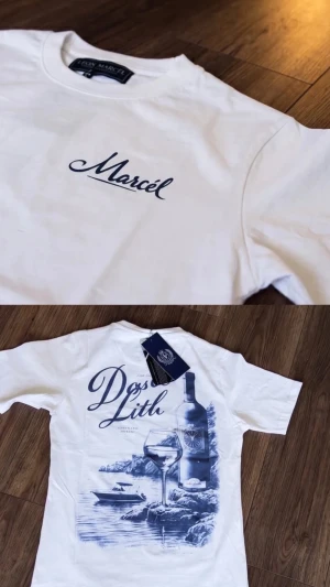 Vit t-shirt från Leon  Marcél - Vit t-shirt från Leon Marcél med blå text 'Marcél' på bröstet och stor grafisk print på ryggen med vinflaska, glas och havsmotiv. Klassisk passform och rund hals. Perfekt för dig som gillar stilrena och unika tryck.