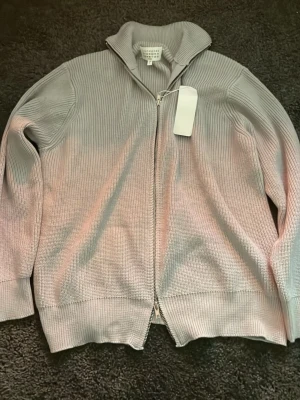 Ljuslila stickad ziptröja Maison Margiela - Snygg ljuslila stickad tröja från Maison Margiela med dragkedja framtill och hög krage. Tröjan har en avslappnad passform och ribbade muddar vid ärmslut och nederkant. Perfekt för lager-på-lager och stilrena outfits.