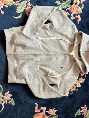 Polo raph lauren sipper - Snygg grå zip-hoodie från Polo Ralph Lauren med klassisk huva och vita dragsnören. Broderad lila logga på bröstet och mjukt, fleecefodrat innerfoder. Perfekt till chill dagar och enkel att matcha med jeans eller joggers.