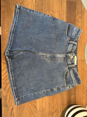 Blå denimshorts med hög midja - Klassiska blå denimshorts med hög midja och raka ben. Shortsen har femficksmodell, bälteshällor och stängs med knapp och dragkedja. Perfekta för sommaren och enkla att matcha med olika toppar. Nypris 899kr