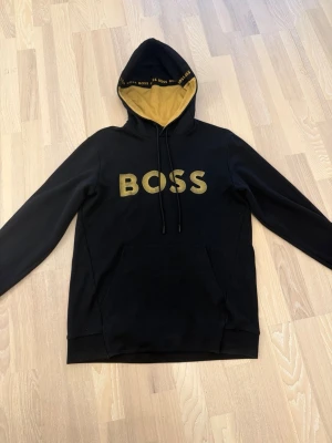 Svart BOSS hoodie - Svart hoodie från BOSS med stor guld broderad logga på bröstet och gul insida i huvan. Tröjan är använd max 5 gånger pga att den alltid har varit för stor på mig.