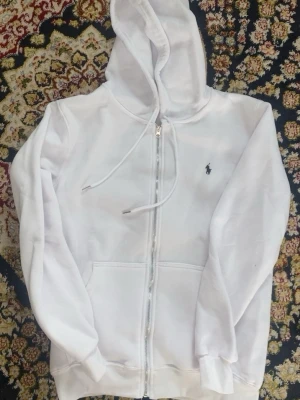 Vit hoodie från Polo Ralph Lauren - Vit hoodie med dragkedja från Polo Ralph Lauren. Klassisk design med huva och snören, två stora fickor framtill och den ikoniska broderade loggan på bröstet. Mjuk bomullsmix som är skön att ha på sig och ribbade muddar vid ärmslut och nederkant.