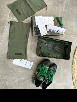 Valentino Garavani gröna sneakers - Säljer ett par snygga sneakers från Valentino Garavani i grönt och svart med coola detaljer och gröna skosnören. Skorna har en låg siluett och är tillverkade i en mix av mocka och läder. Perfekta för dig som vill sticka ut med exklusiva och trendiga sneakers.