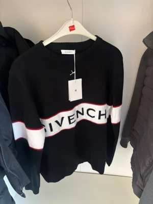 Svart stickad tröja från Givenchy - Snygg svart stickad tröja från Givenchy med vit och röd rand över bröstet och armarna. Stor Givenchy-logga framtill. Rund halsringning och ribbade muddar. Perfekt för dig som gillar streetwear och exklusiva märken. Passar både storlek L och M. Passar de som har L som vill ha den som en smäck på och passar de som har m som vill ha det lite luftigare men ändå sitter bra