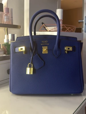 Blå Birkin-liknande handväska 25 cm - Säljer en ikonisk blå handväska i grained läder med guldfärgade metalldetaljer. Helt Oanvänd då jag inte gillade färgen särskilt mycket.