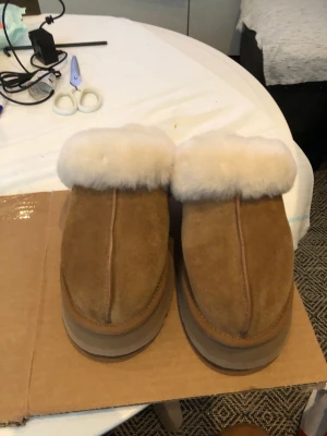Bruna fårskinnstofflor från UGG - Mysiga tofflor från UGG i brunt mockaskinn med fluffig vit fårskinnspäls runt kanten och inuti. Klassisk slip-in modell med rundad tå och rejäl sula. Perfekta för att hålla fötterna varma hemma.