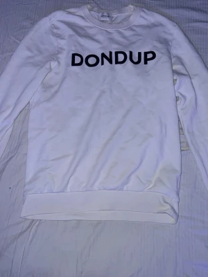 Vit sweatshirt från Dondup - Säljer en stilren vit sweatshirt från Dondup med svart logotyptryck på bröstet. Tröjan har rund halsringning och ribbade muddar vid ärmslut och nederkant. Den har små skador av tvätten på bokstäverna som syns på bild 3. Hör av vid frågor pris kan diskuteras!