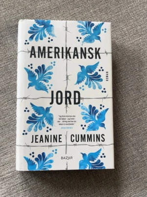 Amerikansk jord av Jeanine Cummins - Romanen 'Amerikansk jord' av Jeanine Cummins har ett vitt omslag med blå fåglar och taggtrådsmotiv. Boken är en internationell bästsäljare och handlar om flykt från kartellvåld i Mexiko. Inbunden och tryckt av Bazar förlag.