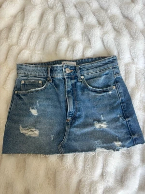 Blå jeanskjol från Zara - Säljer en blå jeanskjol från Zara med slitna detaljer och rå kant nertill. Kjolen har klassisk femficksdesign och knappgylf. Perfekt för dig som gillar en avslappnad och trendig stil.