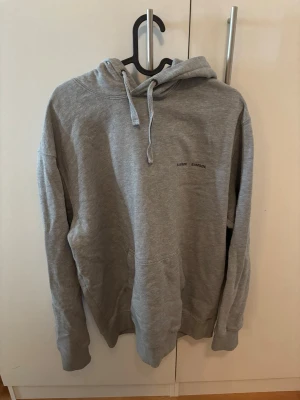 Grå hoodie från Samsøe Samsøe - Snygg grå hoodie från Samsøe Samsøe med minimalistisk logga på bröstet. Klassisk modell med huva och dragsnören samt en stor magficka. Tillverkad i mjukt material som är skönt att ha på sig. Perfekt för en avslappnad stil.