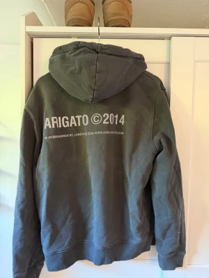 Grå hoodie från Axel Arigato - Snygg grå hoodie från Axel Arigato med tryck 'ARIGATO ©2014' på både bröstet och ryggen. Klassisk känguruficka framtill och stor huva. Perfekt för en avslappnad streetwear-look.