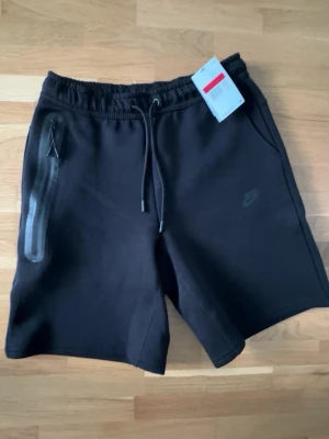 Svarta shorts från Nike med snörning - Svarta shorts från Nike med elastisk midja och snörning. Klassisk design med diskret Nike-logga på vänster ben och dragkedjeficka på höger sida. Perfekta för en sportig och avslappnad stil.