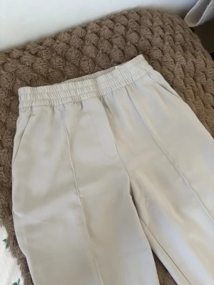 Beiga kostymbyxor med resårmidja - Säljer ett par beiga byxor med vida ben och resår i midjan. Byxorna har pressveck framtill och sidofickor. Perfekta för en avslappnad men ändå stilren look. Materialet känns mjukt och följsamt.