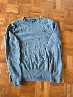 Grå stickad tröja Polo Ralph Lauren - Snygg grå stickad tröja från Polo Ralph Lauren med rund halsringning och diskret broderad logga på bröstet. Tröjan har ribbade muddar vid ärmslut och nederkant. Perfekt till jeans eller chinos för en clean look.