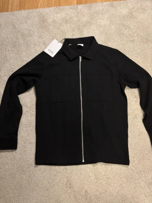 Helt ny svart overshirt från Selected Homme - Helt ny stilren svart overshirt från Selected Homme med rak passform, två bröstfickor och hel dragkedja framtill. Klassisk krage och långa ärmar. Perfekt att bära som lager över en t-shirt. Materialet känns mjukt och har en lätt struktur.