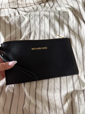 Svart kuvertväska Michael Kors - Snygg svart kuvertväska från Michael Kors i äkta läder med guldfärgade detaljer och dragkedja. Väskan har ett stilrent och klassiskt utseende med logga framtill och praktisk handledsrem. Perfekt för att bära det viktigaste när du är på språng.