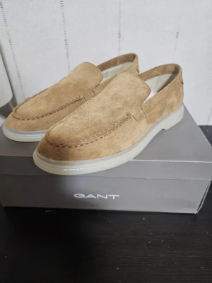 Beige mockaloafers från Gant - Snygga loafers i beige mocka från Gant med klassisk design och rundad tå. Skorna har en vit, robust sula som ger en modern touch. Perfekta för dig som gillar stilrena och tidlösa skor i mjukt material.