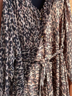 Leopardmönstrad långklänning med volang - Säljer en långklänning med leopardmönster och volangdetaljer vid v-ringningen. Klänningen har långa ärmar, knappar framtill och ett matchande midjebälte. Materialet är tunt och flowy, perfekt för dig som vill sticka ut.Det står 3XL 48 men den är som L