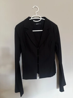 Svart kavaj från Gina Tricot - Stilren svart kavaj från Gina Tricot med klassisk krage och smal passform. Kavajen har diskreta hyskor som knäppning framtill och långa ärmar. Perfekt för en snygg och uppklädd look. Aldrig använd