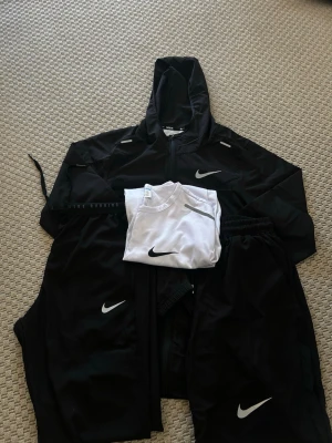 Nike milner - Snyggt svart träningsset från Nike med jacka med huva, byxor och en vit t-shirt. Jackan har dragkedja och reflexdetaljer, byxorna har resår i midjan och Nike-logga på benet. T-shirten är vit med svart Nike-logga. Perfekt för löpning eller gym.
