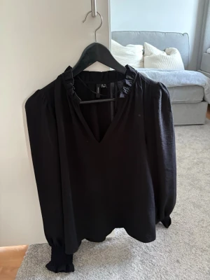 Svart blus - Stilren svart blus från Vero Moda med v-ringning och volangdetalj vid kragen. Blusen har långa ärmar med resår. Tillverkad i ett mjukt, glansigt material som ger en elegant känsla.