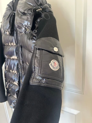 Svart dunjacka från Moncler - Snygg svart pufferjacka från Moncler med glansig kropp och stickade ärmar. Jackan har dragkedja framtill, ficka med Moncler-logga på ärmen och huva. Perfekt för dig som vill ha en trendig och bekväm jacka.