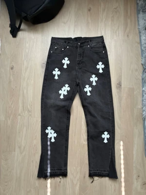 Flared ChromeHearts Jeans - Unika svarta jeans med flera vita korsapplikationer framtill. Raka ben med råa, fransiga benslut och slits på ena benet för extra edge. Jeansen har klassisk femficksdesign och är tillverkade i kraftigt denimtyg. Perfekta för dig som gillar streetwear och statement-plagg.