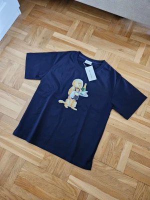 Mörkblå t-shirt Drôle de Monsieur - Snygg mörkblå t-shirt från Drôle de Monsieur med ett stort tryck framtill av en hund som servitör med bricka, champagneflaska och glas. Klassisk passform och rund hals. Perfekt för dig som gillar unika prints och streetwear vibes.