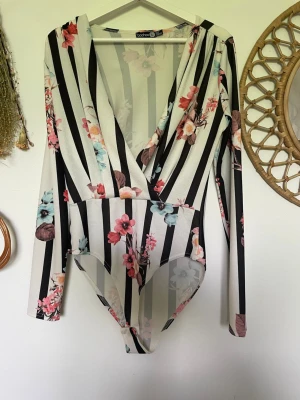 Randig blommig omlottbody Boohoo - Säljer en långärmad body från Boohoo med svartvita ränder och blommigt mönster i rosa, blått och beige. Omlott framtill med djup v-ringning.