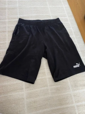 Svarta shorts från Puma - Säljer ett par klassiska svarta shorts från Puma med elastisk midja och logga på vänster ben. Perfekta för träning eller chill, tillverkade i mjuk bomull för skön känsla. Enkla att matcha och har fickor på sidorna.