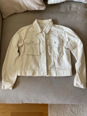 Vit jeansjacka från NLY Trend - Snygg vit jeansjacka från NLY Trend med raka linjer och två stora bröstfickor med lock. Klassisk krage och knäppning framtill. Perfekt att slänga över en t-shirt för en clean och fräsch look.