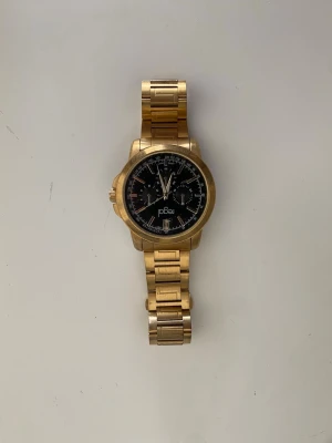 Guldig klocka från Póal med svart urtavla - Stilren klocka från Póal i guldigt metallarmband och svart urtavla med vita detaljer. Klockan har klassisk rund form och robust länkarmband som ger en lyxig känsla. Perfekt accessoar för att lyfta din outfit.