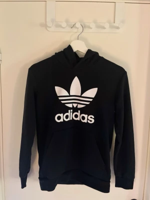 Svart hoodie från Adidas med logga - Snygg svart hoodie från Adidas med stor vit Trefoil-logga och text på bröstet. Klassisk känguruficka framtill och huva. Tillverkad i mjukt bomullsmaterial som är skönt att ha på sig. Perfekt för en avslappnad streetwear-look.