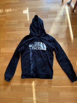 Svart hoodie från The North Face - Svart hoodie från The North Face med stor vit logga på bröstet. Klassisk modell med huva och dragsko samt känguruficka framtill. Har en reva i resåret baktill, syns på sista bilden. 