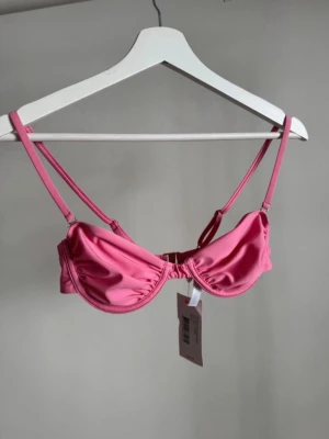Rosa bikiniöverdel med bygel - Snygg rosa bikiniöverdel med bygel och justerbara axelband. Modellen har en klassisk balconette-form och stängs med spänne i ryggen. Perfekt för strandhäng och poolparty. Materialet är mjukt och följsamt för bästa komfort. Aldrig använd!!