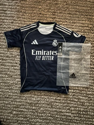 Mörkblå Real Madrid matchtröja Adidas - Snygg mörkblå Real Madrid matchtröja från Adidas med vita detaljer och klubbmärke på bröstet. Tröjan har vita ränder på axlarna, trycket 'Emirates Fly Better' på framsidan och är tillverkad i ett lätt, andningsbart funktionsmaterial. Perfekt för fotbollsträning eller match.