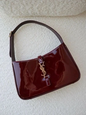 Vinröd axelväska från Saint Laurent - Snygg vinröd axelväska från Saint Laurent i glansigt lackläder. Väskan har en klassisk rektangulär form, justerbart axelband och guldfärgad YSL-logga framtill. Perfekt för dig som vill ha en elegant och trendig accessoar med lyxig känsla.