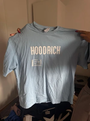 Blå HOODRICH t-shirt med tryck - Snygg ljusblå t-shirt från HOODRICH med vitt tryck både fram och bak. Framsidan har stor logga och text, baksidan har flera grafiska tryck och texten 'FROM NOTHING TO SOMETHING'. Klassisk passform och rund hals. Perfekt för en streetwear-look.