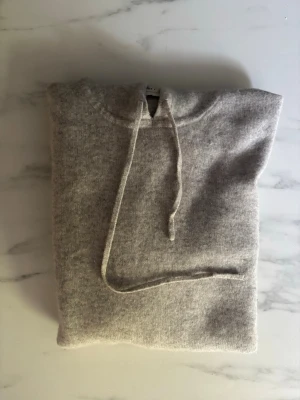 Grå hoodie från Soft Goat i cashmere - Säljer en grå hoodie från Soft Goat i mjuk cashmere. Hoodien har huva med dragsko och långa ärmar. Perfekt för dig som gillar stilrena och bekväma plagg. Ribbstickade muddar vid ärmslut och nederkant. Enkel och klassisk design.