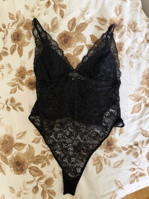 Svart spetsbody med v-ringning - Snygg svart body i genombruten spets med djup v-ringning och smala axelband. Bodyn har blommigt spetsmönster och transparenta partier som ger en elegant look. Perfekt för dig som vill ha något extra under kläderna.