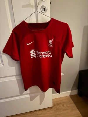 Liverpool matchtröja Nike röd - Röd Liverpool FC matchtröja från Nike med korta ärmar. Tryck med klubbmärke, sponsorlogga och spelarnamn 'Virgil' samt nummer 4 på ryggen. Perfekt för fotbollsträning eller att bära på matchdag. Klassisk passform och sportig design.