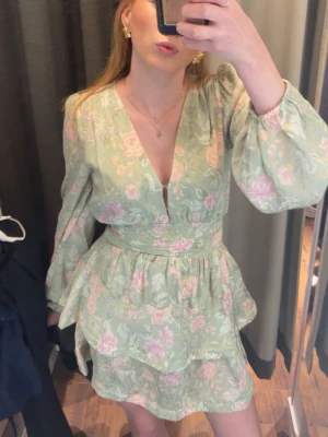 Grön blommig kortklänning med volang - Söt kortklänning i ljusgrönt med blommigt mönster i rosa och vitt. Klänningen har v-ringning, långa puffärmar och markerad midja med volangdetaljer i kjolen. Perfekt för somriga dagar och midsommarfirande. Bara använt en gång 