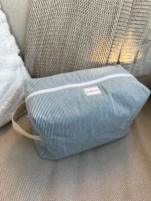 Randig necessär från Emelo - Snygg necessär från Emelo i blåvitt randigt jeanstyg. Rektangulär form med vit dragkedja och praktiskt handtag i beige tyg på sidan. Perfekt för att organisera småsaker eller smink. Tillverkad i jeans tyg. Innerfack finns. Mått ca 26x17x12cm