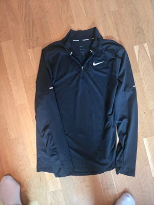 Svart långärmad träningströja Nike - Svart långärmad träningströja från Nike med halv dragkedja och vit logga på bröstet. Tröjan har en sportig passform och är tillverkad i ett lätt och stretchigt material som passar perfekt för löpning eller gym. Snygga detaljer vid ärmarna.