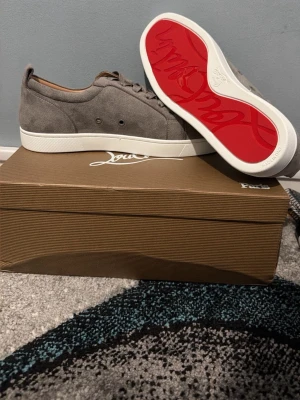 Gråa Louboutin sneakers i mocka - Snygga gråa sneakers från Christian Louboutin i mjuk mocka med klassisk röd sula och vita kanter. Lågt skaft och stilren design med ton-i-ton snörning och diskreta detaljer. Perfekta för dig som vill ha något exklusivt och trendigt.