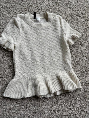 Beige stickad topp med volang - Söt beige stickad topp med korta ärmar och volang nedtill. Tröjan har ett diskret mönster och ribbstickade detaljer vid hals, ärmslut och nederkant. Perfekt för en trendig och avslappnad look.