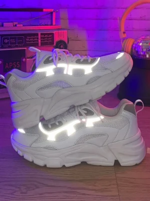 Chunky vita sneakers med reflexdetaljer - Säljer ett par chunky vita sneakers med feta sulor och reflekterande detaljer som lyser upp i ljus. Skorna har meshpaneler för extra andningsförmåga och cool look. Perfekta för dig som vill sticka ut och gillar streetwear-vibbar. Sköna att gå i och riktigt trendiga.