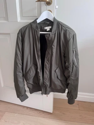 Olivgrön bomberjacka från H&M - Snygg olivgrön bomberjacka från H&M med klassisk ribbad krage, muddar och dragkedja framtill. Jackan har två snedställda fickor med lock och en ärmficka med dragkedja. Tillverkad i ett glansigt syntetmaterial som ger en modern look. Kom privat för fler bilder💚
