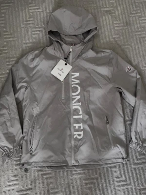 Grå vindjacka från Moncler - Snygg grå vindjacka från Moncler med huva och dragkedja framtill. Jackan har stora vita Moncler-bokstäver längs framsidan, två dragkedjefickor och elastiska muddar vid ärmsluten. Tillverkad i lätt, vindtätt material och har Moncler-logga på ärmen.
