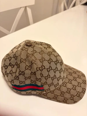 GG Keps - Snygg keps från Gg i beige med klassiskt GG-monogram över hela kepsen. Har en ikonisk röd och grön rand på sidan och justerbar rem baktill. Materialet är canvas med svart insida och märkta detaljer. Perfekt för dig som vill sticka ut med lyxig streetstyle.